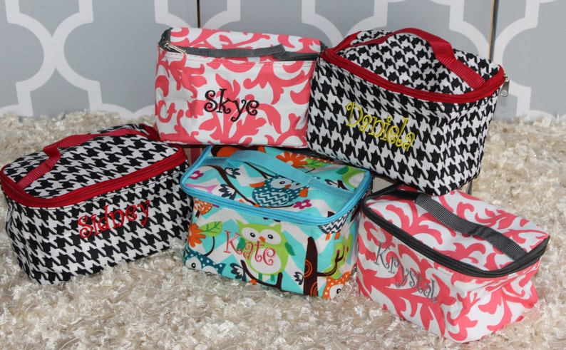 Op de afbeelding: Vijf gepersonaliseerde lunchtassen met verschillende patronen en kleuren. De tassen hebben namen geborduurd in verschillende kleuren. Patronen zijn onder meer zwart-wit hondenpoot, koraalroze bloemen en een blauw-groen uilontwerp. Elke tas heeft een handvat.