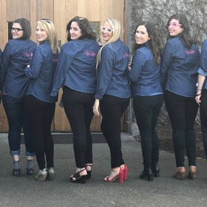 May include: A group of nine people wearing denim shirts with the text "She Said Yes" embroidered on the back. They are standing in front of a wooden door and a stone wall.
