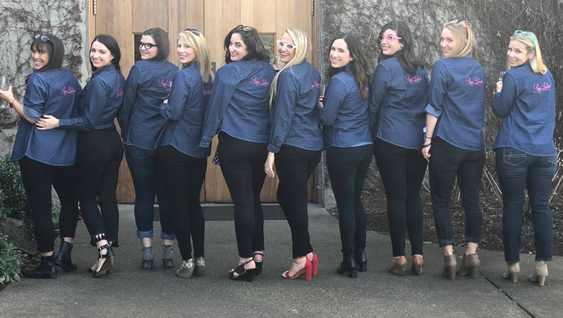 May include: A group of nine people wearing denim shirts with the text "She Said Yes" embroidered on the back. They are standing in front of a wooden door and a stone wall.