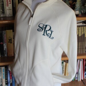 May include: Cream-colored fleece jacket with a full zipper and a stand-up collar. The jacket features a teal monogram of the letters "SRL" on the left chest. The jacket is displayed on a mannequin and is surrounded by a bookshelf filled with books.