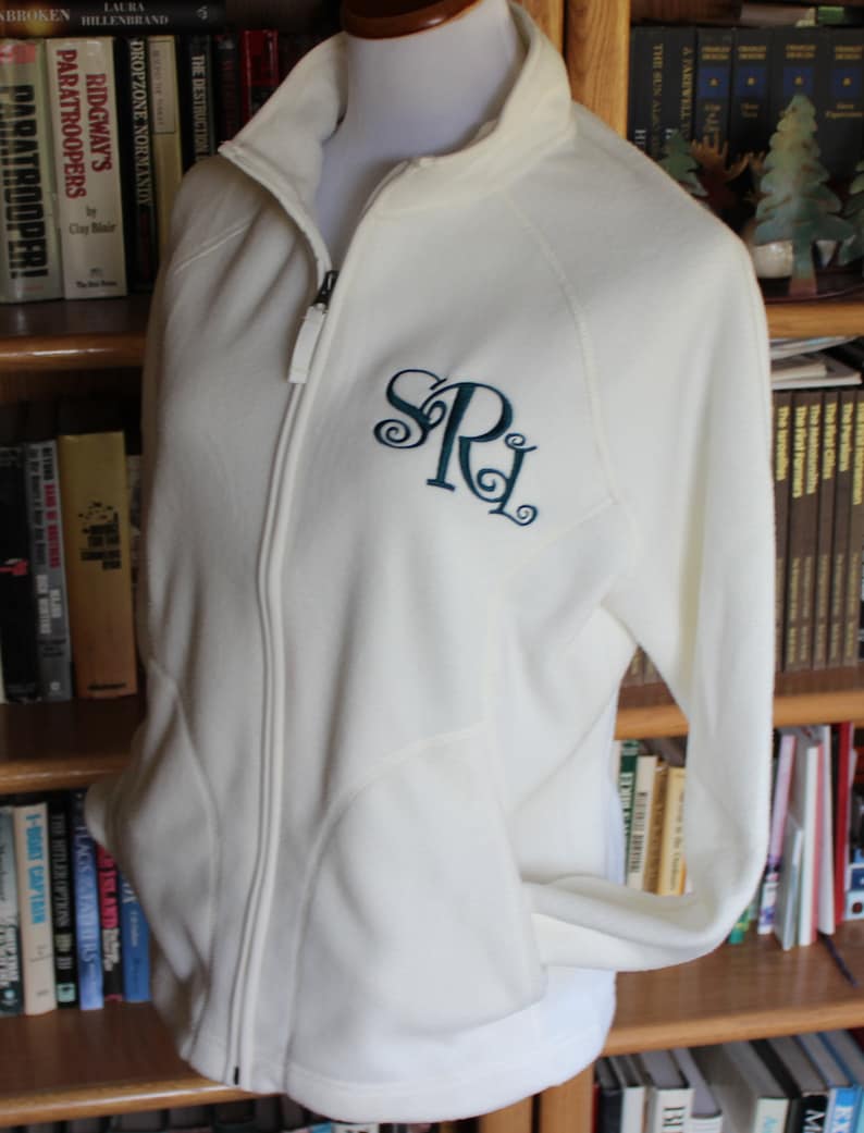 May include: Cream-colored fleece jacket with a full zipper and a stand-up collar. The jacket features a teal monogram of the letters "SRL" on the left chest. The jacket is displayed on a mannequin and is surrounded by a bookshelf filled with books.