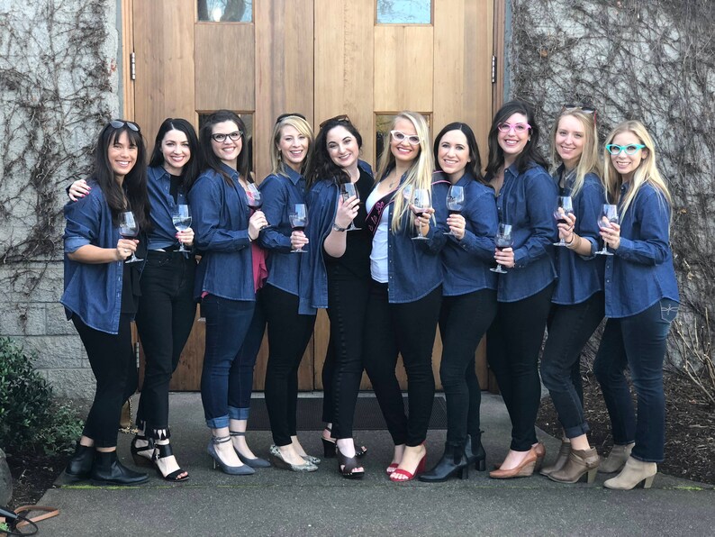 May include: A group of women wearing denim shirts and black trousers are holding wine glasses. They are standing in front of a wooden door with a stone wall behind them.