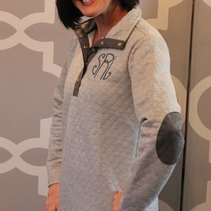 May include: A gray quilted pullover with a gray patch on the elbow and a monogrammed "SR" on the chest. The pullover has a snap button closure at the neck.