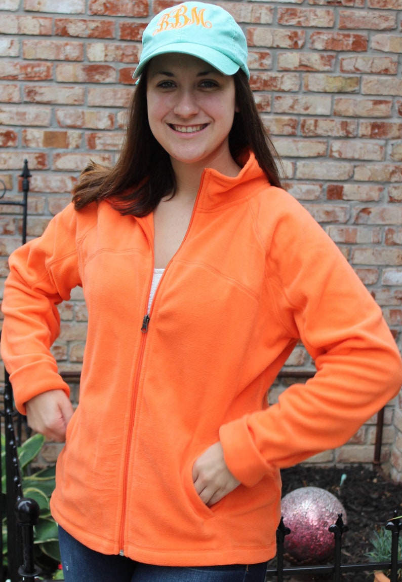 May include: An orange fleece jacket with a full zipper and side pockets. The person is wearing a light blue baseball cap with the embroidered letters "BBM". The background features a brick wall and outdoor foliage.