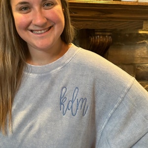 May include: A light blue, striped, long-sleeved sweatshirt with the word "kdm" embroidered on the front in blue thread.