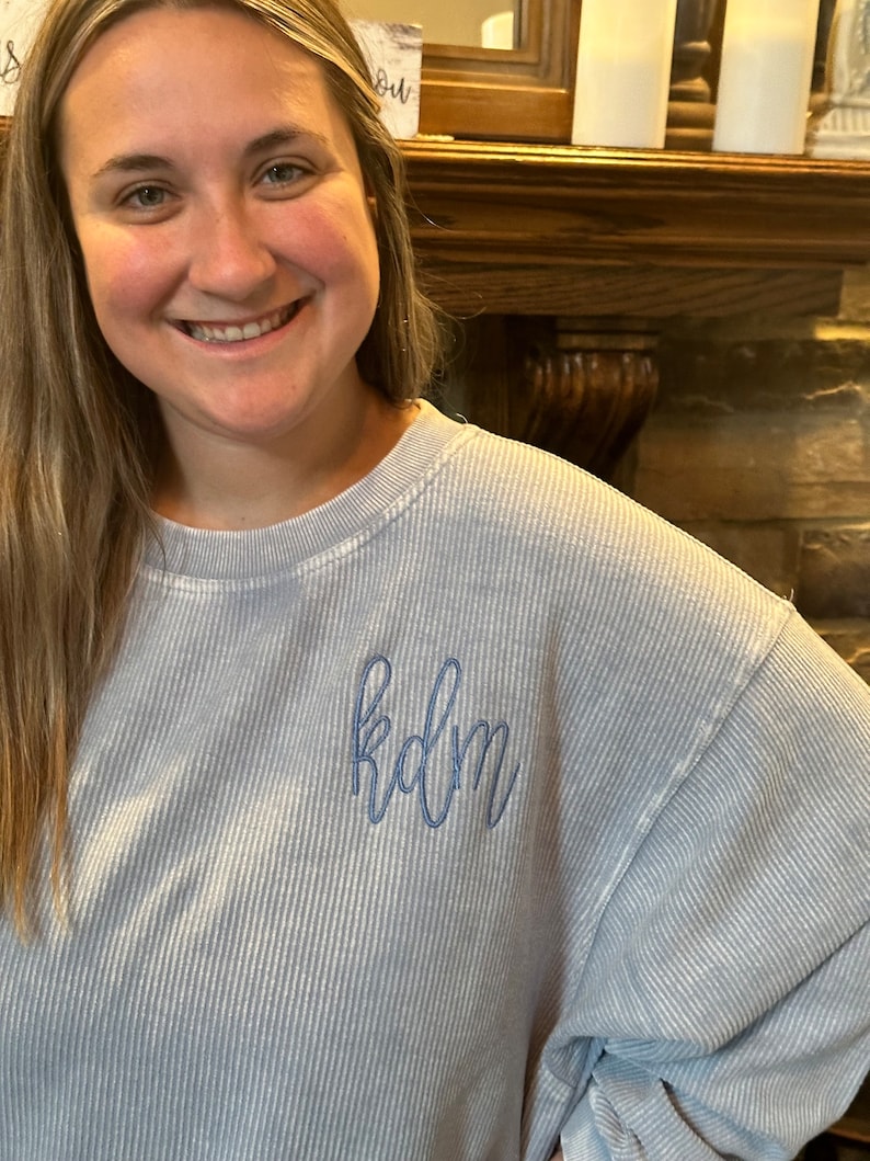 May include: A light blue, striped, long-sleeved sweatshirt with the word "kdm" embroidered on the front in blue thread.
