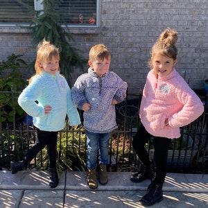 May include: Three children wearing fleece pullovers. The child on the left is wearing a light blue pullover, the child in the middle is wearing a gray pullover, and the child on the right is wearing a pink pullover.