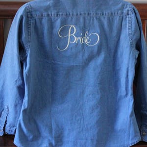 K&ouml;nnte beinhalten: Ein blaues Jeanshemd mit dem Wort "Bride" in wei&szlig;er Schrift auf der R&uuml;ckseite gestickt.