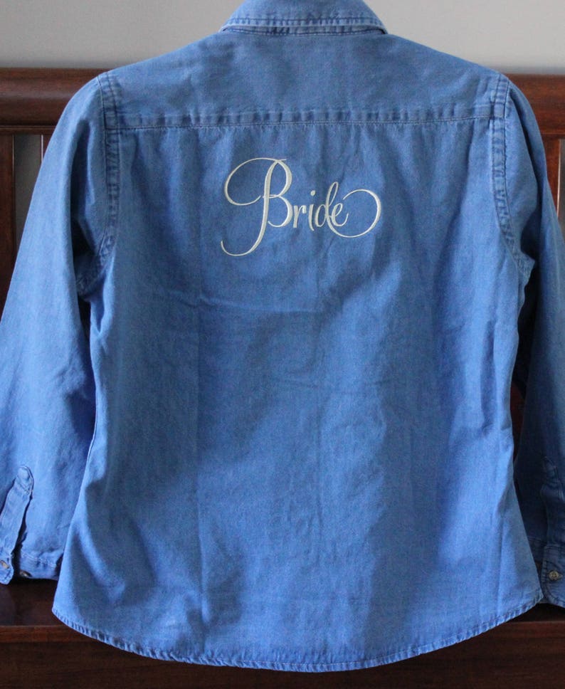 K&ouml;nnte beinhalten: Ein blaues Jeanshemd mit dem Wort "Bride" in wei&szlig;er Schrift auf der R&uuml;ckseite gestickt.