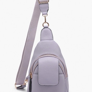 May include: A light purple sling bag with a matching adjustable shoulder strap. The bag features a front pocket, a main compartment with a zipper, and gold-colored hardware. The bag is made of a smooth, leather-like material.