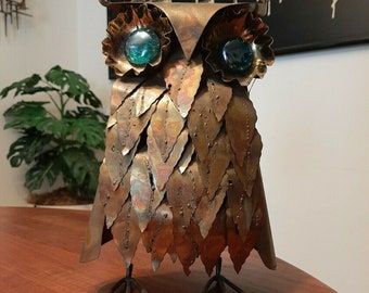 May include: A vintage metal owl sculpture with a copper finish. The owl features leaf-shaped details, teal glass eyes, and a decorative loop above the head. The sculpture is standing on a wooden surface.