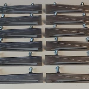 May include: A collection of rectangular cabinet pulls in various shades of gray and bronze. Each pull has a linear design and is mounted with a silver-colored screw. The pulls are arranged in rows on a light-colored surface.