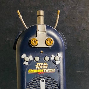 Puede incluir: Un walkie talkie azul Star Wars CommTech con detalles dorados y un botón rojo. El walkie talkie tiene una superficie texturizada y una antena plateada.