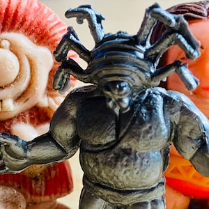 May include: Three plastic figurines, one red and white, one grey with multiple arms, and one pink with brown hair. The grey figurine has a striped head and appears to be a monster.