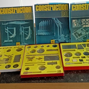 Könnte beinhalten: Drei blaue Boxen mit dem Wort "Construction" und einer Nummer auf jeder Box. Die Boxen sind mit "Construction 100", "Construction 101" und "Construction 200" beschriftet. Jede Box enthält einen Satz kleiner Metallteile zum Bau von Modellen.