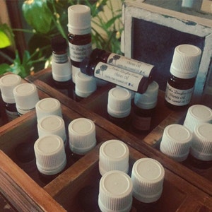 Peut inclure: Une boîte en bois remplie de petits flacons en verre avec des couvercles blancs. Les flacons contiennent des huiles essentielles. Les étiquettes sur les flacons indiquent "Thieves Oil" et "100% Pure Essential Oil".