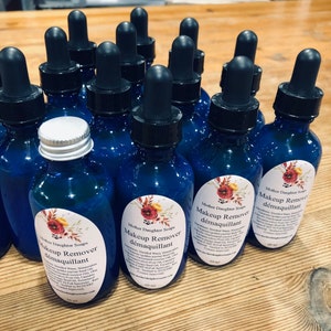 May include: A close-up of several blue glass bottles with dropper tops, filled with a clear liquid. Each bottle has a white label with the text "Mother Daughter Soaps" and "Makeup Remover d&eacute;maquillant" in black lettering. The labels also feature a floral design with red, yellow, and green colours.