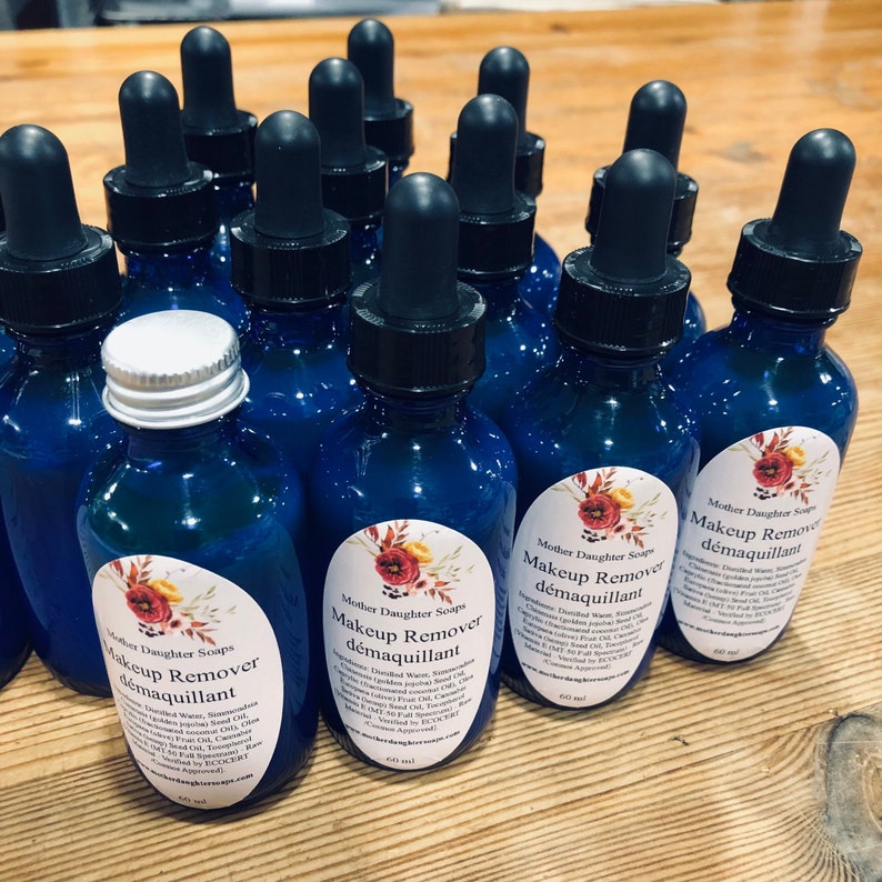 May include: A close-up of several blue glass bottles with dropper tops, filled with a clear liquid. Each bottle has a white label with the text "Mother Daughter Soaps" and "Makeup Remover d&eacute;maquillant" in black lettering. The labels also feature a floral design with red, yellow, and green colours.