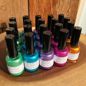May include: A collection of 20 small bottles of nail polish in various colours, including green, blue, purple, pink, and gold. The bottles are arranged in a semi-circle on a wooden surface. Each bottle has a black cap and a label that reads "Vegan Cruelty Free & 10-Free Nail Polish Vernis ongles".
