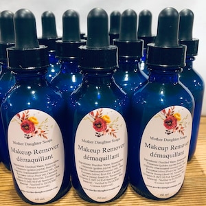 May include: A group of 12 blue glass bottles with dropper tops, each containing 60 ml of makeup remover. The bottles have labels with the text "Mother Daughter Soaps" and "Makeup Remover d&eacute;maquillant" in white lettering. The labels also list the ingredients of the makeup remover.
