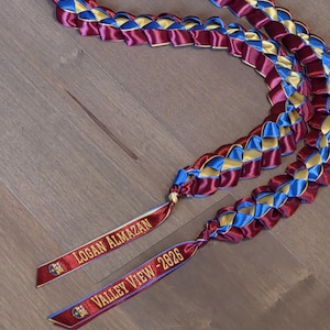 May include: A braided graduation lei in maroon, blue, and gold. The lei features two ribbons with the text "LOGAN ALMAZAN" and "VALLEY VIEW - 2026". The lei is made of satin ribbon and is displayed on a wooden surface.