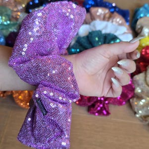 May include: A bright purple fabric hair scrunchie covered in small, reflective sequins. The scrunchie features a gathered, elasticized design and includes a small, rectangular black fabric tag attached to the side of the purple material.