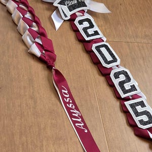 May include: Two graduation leis. One is a maroon and white braided design with a maroon ribbon that says "Alyssa". The other lei has the text "Class of" and "2025" in white and black.