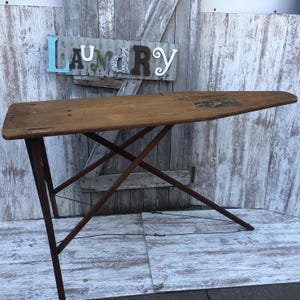 K&ouml;nnte beinhalten: Ein vintage B&uuml;gelbrett aus Holz mit einer braunen Holzplatte und einem schwarzen Metallrahmen. Das Brett ist zusammengeklappt und steht auf einem wei&szlig;en Holzboden. Das Wort "Laundry" ist in bunten Buchstaben auf einem Metallschild &uuml;ber dem Brett geschrieben.
