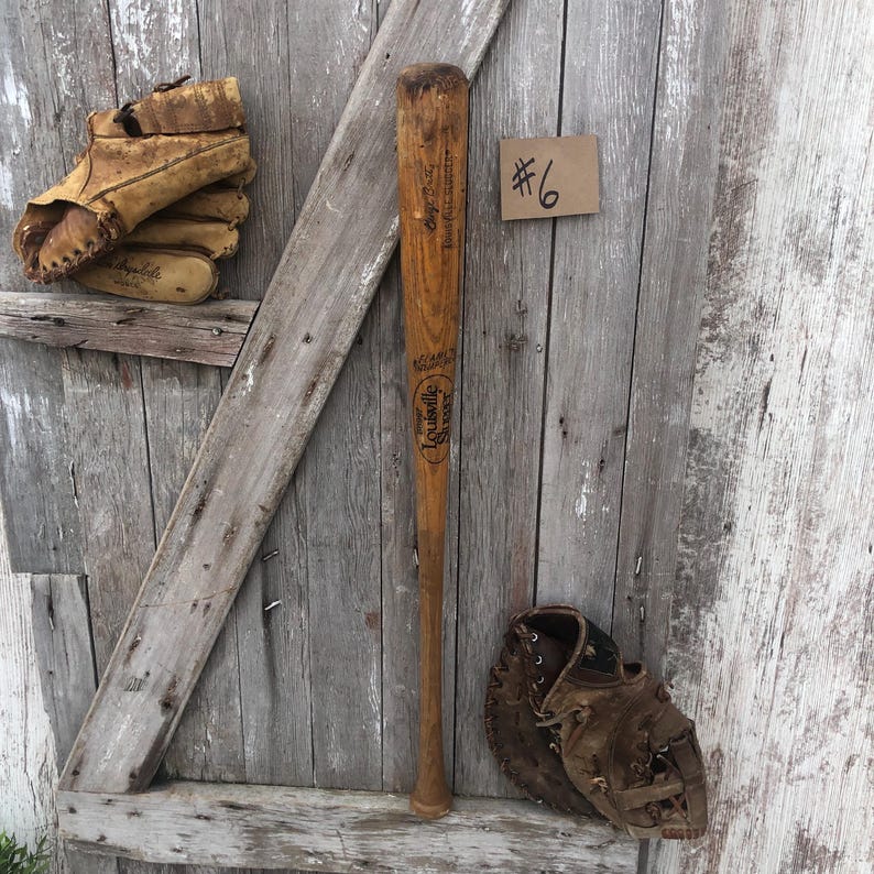 Puede incluir: Un bate de b&eacute;isbol vintage y dos guantes de cuero desgastados se exhiben sobre un fondo de madera envejecida. El bate, marcado "Louisville Slugger", est&aacute; colocado verticalmente. Una peque&ntilde;a etiqueta dice "#6".