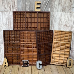 May include: Five rectangular wooden printer trays with multiple small compartments of varying sizes arranged in a grid pattern. The trays feature different wood tones ranging from light to dark brown. The letters A, B, C, D, and E are placed in front.
