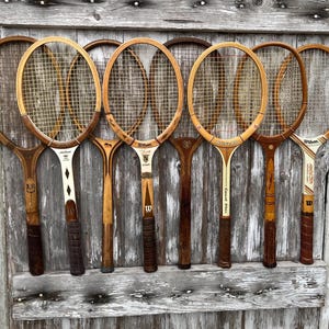 May include: A collection of seven vintage wooden tennis rackets arranged in a row. The rackets feature various wood tones, nylon string patterns, and branded handles including Wilson and Dunlop, with visible text markings like Two Star and Victory.
