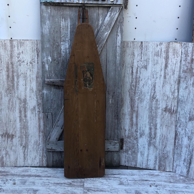 K&ouml;nnte beinhalten: Ein antikes B&uuml;gelbrett aus Holz mit einem Etikett mit der Aufschrift "The Perfect Ironing Board". Das Brett ist braun und hat eine leicht verwitterte Optik.