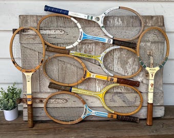 Op de afbeelding: Een verzameling van zeven vintage houten tennisrackets met verschillende merknamen, waaronder Wilson, Dunlop en Performer. De rackets hebben diverse kleurenschema's zoals geel, blauw, wit en natuurlijke houttinten met nylon snaren.