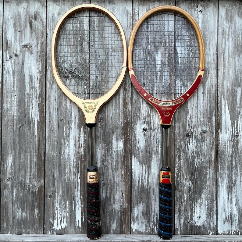 May include: Two vintage wooden tennis rackets with strung heads. The left racket features a light wood frame with a gold Tourney logo. The right racket has a red and wood frame with Tourney-I Super text and a crown graphic. Both have wrapped handles.