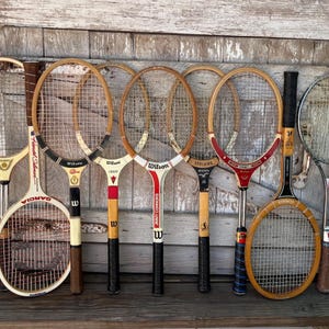 May include: A collection of eight vintage tennis rackets featuring various wooden and metal frames with nylon string patterns. The rackets display diverse brand names including Wilson, Garcia, and Spalding, with distinct color accents in red, black, and white.