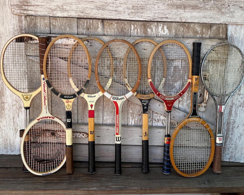 May include: A collection of eight vintage tennis rackets featuring various wooden and metal frames with nylon string patterns. The rackets display diverse brand names including Wilson, Garcia, and Spalding, with distinct color accents in red, black, and white.