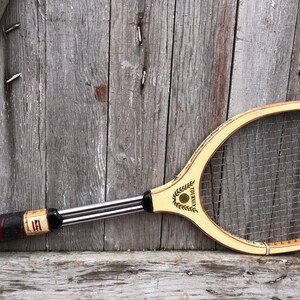 May include: A vintage wooden tennis racket featuring a light-colored frame with a black and red grip. The throat of the racket displays the text TOURNEY with a laurel wreath graphic. It has a metal shaft and a standard nylon string pattern.