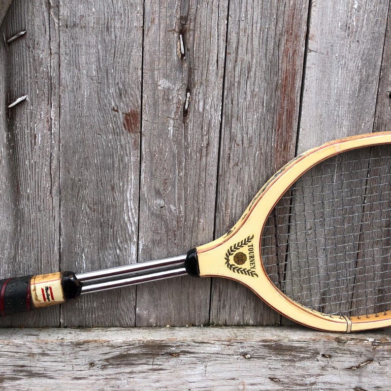 May include: A vintage wooden tennis racket featuring a light-colored frame with a black and red grip. The throat of the racket displays the text TOURNEY with a laurel wreath graphic. It has a metal shaft and a standard nylon string pattern.