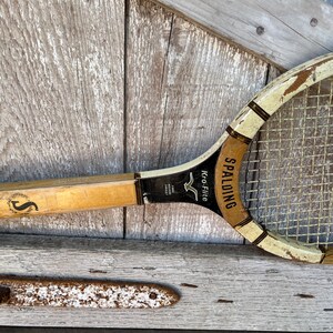 May include: A vintage wooden tennis racket with a cream and black frame, featuring the text Spalding Kro-Flite and a letter S logo on the handle. The racket has a woven string pattern and shows signs of wear on the painted wooden surface.
