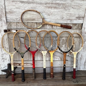 May include: A collection of eight vintage wooden tennis rackets arranged in a row. The rackets feature various natural wood tones, red, black, and white paint accents, and woven nylon strings. Each racket has a distinct handle wrap and frame design.