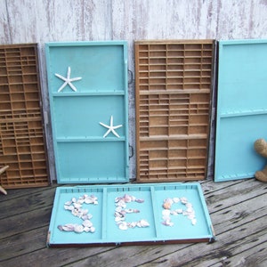 May include: Five wooden trays, two painted light blue, two painted brown, and one painted light blue with the word "SEA" spelled out in seashells. The trays have small compartments and are arranged in a row on a wooden floor.