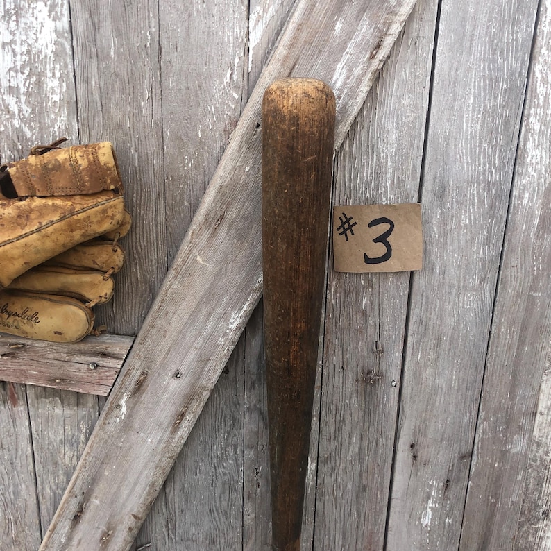 Puede incluir: Un bate de b&eacute;isbol de madera vintage, de color marr&oacute;n medio, con la parte superior redondeada y el cuerpo c&oacute;nico. El bate muestra signos de desgaste. Un guante de b&eacute;isbol de color beige es visible en el fondo. Una peque&ntilde;a etiqueta con el n&uacute;mero "3" est&aacute; adjunta.