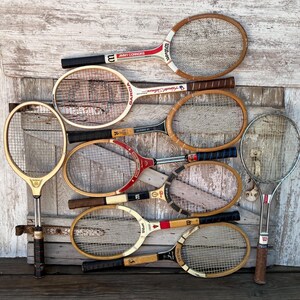 May include: A collection of nine vintage tennis rackets featuring wooden frames with various brand names like Wilson, Garcia, and Spalding, along with one modern metal racket, all arranged together in a group with diverse grip colors and string patterns.