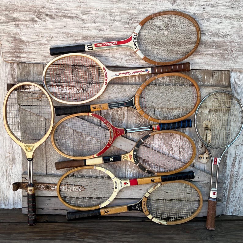 May include: A collection of nine vintage tennis rackets featuring wooden frames with various brand names like Wilson, Garcia, and Spalding, along with one modern metal racket, all arranged together in a group with diverse grip colors and string patterns.