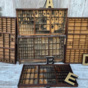 May include: A collection of five rectangular wooden printer's trays featuring numerous small compartments of varying sizes. The trays have a dark wood finish with worn, light-colored interior bases. Decorative letters A, B, C, D, and E are included.
