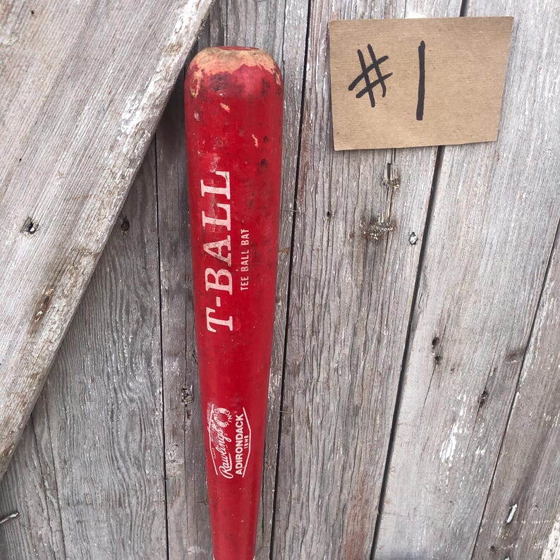 Puede incluir: Un bate de T-ball rojo con letras blancas que dicen "T-BALL TEE BALL BAT". El bate tiene un logotipo de Rawlings Adirondack y una etiqueta de cart&oacute;n con el n&uacute;mero "1". Se apoya contra un fondo de madera desgastada.