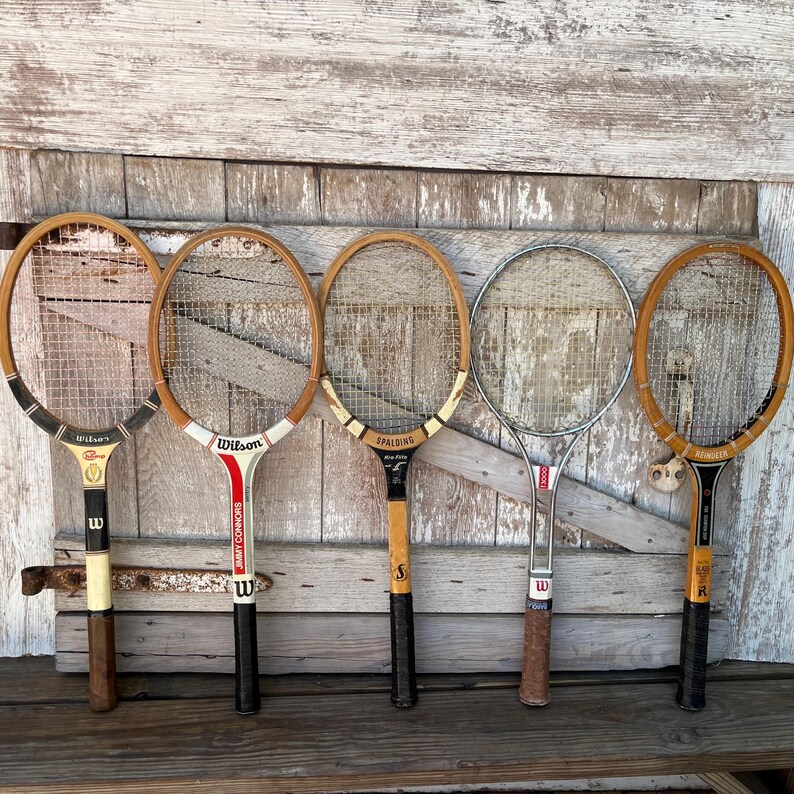 May include: Five vintage tennis rackets arranged in a row. The collection includes three wooden rackets with varied branding like Wilson and Spalding, one metal Wilson T3000 racket, and one wooden racket with black and tan accents and white strings.