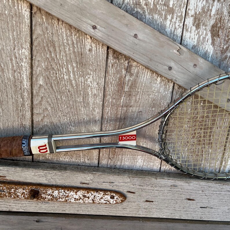 May include: A vintage Wilson T3000 tennis racket featuring a polished chrome metal frame, a brown leather-wrapped handle, and white nylon strings. The throat of the racket displays the Wilson logo and the model name T3000 in red and white text.