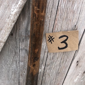 Puede incluir: Primer plano de un poste de madera con acabado marr&oacute;n oscuro. Un peque&ntilde;o trozo rectangular de cart&oacute;n con el n&uacute;mero "3" escrito con tinta negra est&aacute; adherido a la superficie de madera.