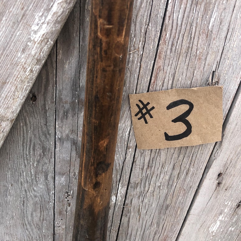 Puede incluir: Primer plano de un poste de madera con acabado marr&oacute;n oscuro. Un peque&ntilde;o trozo rectangular de cart&oacute;n con el n&uacute;mero "3" escrito con tinta negra est&aacute; adherido a la superficie de madera.